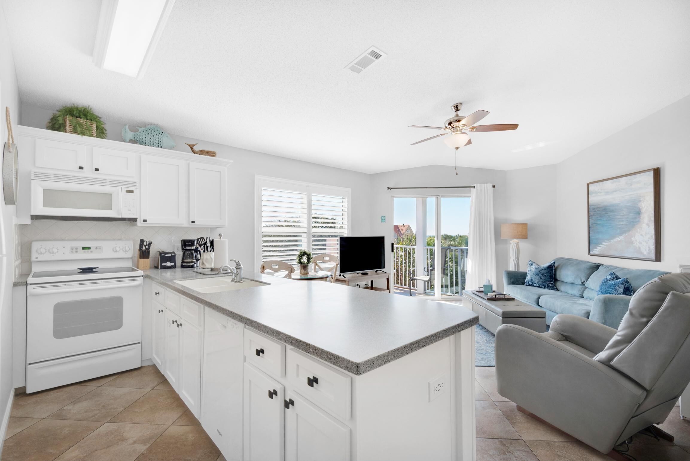 A modern kitchen with white cabinets, a built-in microwave, and a large island. There is a dining area with a table and chairs, and a living room with a gray sofa and a ceiling fan.