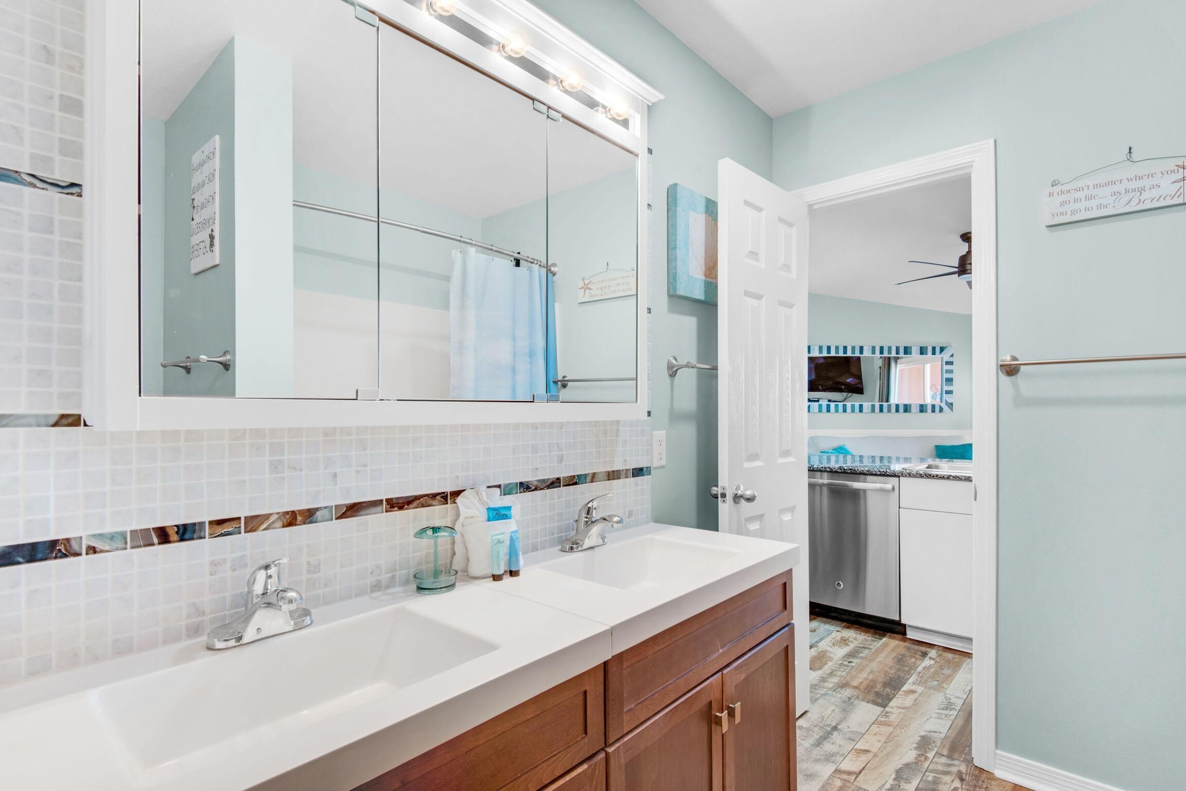 A bathroom with a double sink, a large mirror with lights, a shower area, and a view into the kitchen.