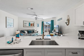 Een moderne keuken met een centraal eiland, een plafondventilator, en uitzicht op de woonkamer met een bank en een bordje met de tekst 'BEACH HOUSE'.