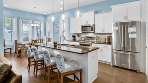 A modern kitchen with a central island, white cabinetry, stainless steel appliances, pendant lighting, and a dining area with patterned chairs. - Fun In The Sun (Miramar Beach)
