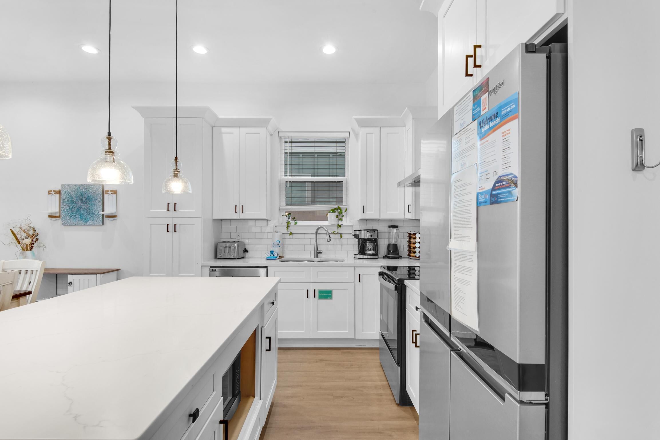 A modern kitchen with white cabinetry, a large island, stainless steel appliances, pendant lighting, and a window with blinds.