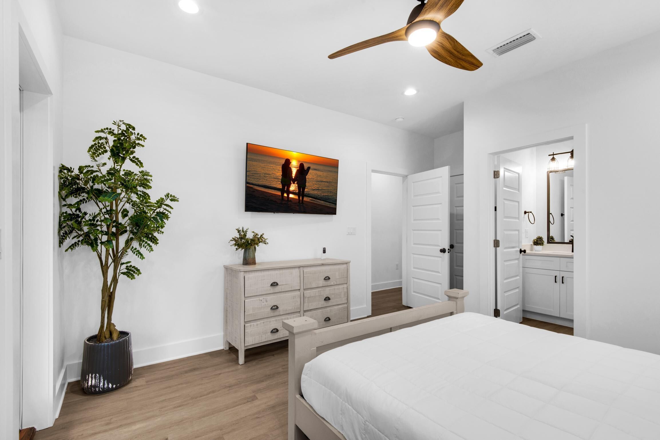 A bedroom with a bed, a dresser, a potted plant, a ceiling fan, and a painting of a sunset.