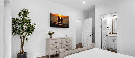 A bedroom with a bed, a dresser, a potted plant, a ceiling fan, and a painting of a sunset.