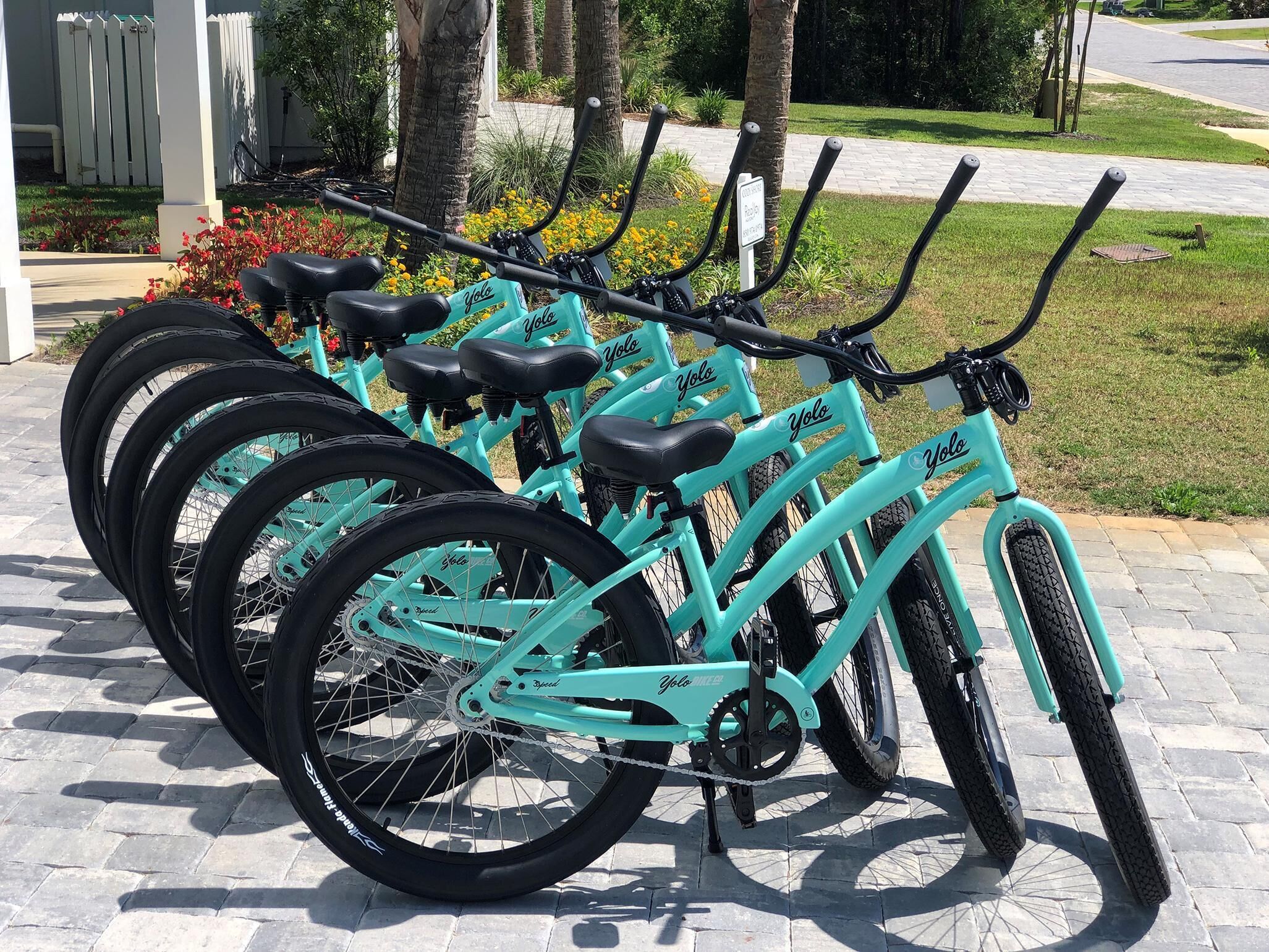 A row of turquoise bicycles parked on a paved area with a grassy background and a white fence.