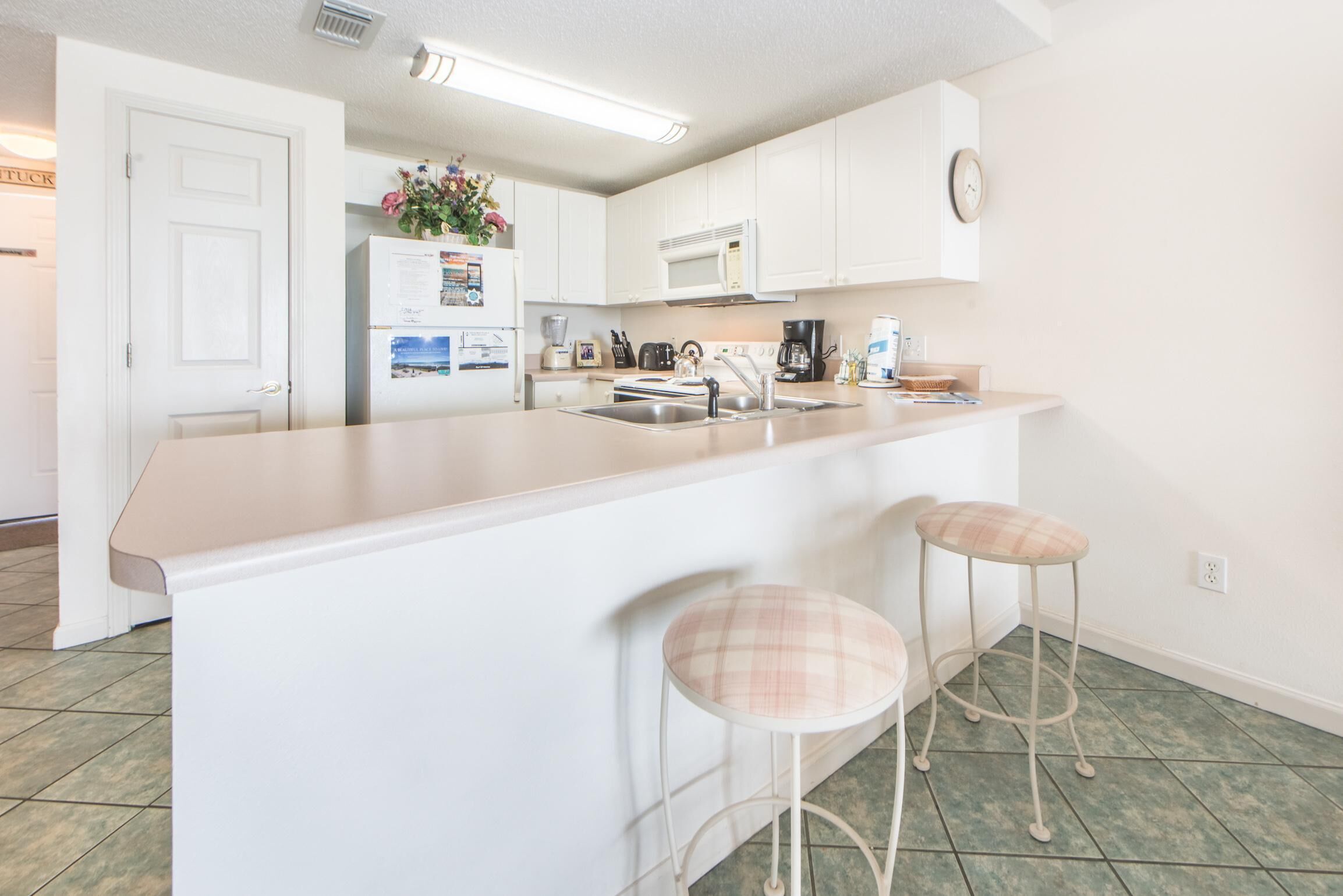 A kitchen with white cabinets, a stainless steel sink, a microwave, and a coffee maker. There are two bar stools with plaid cushions.