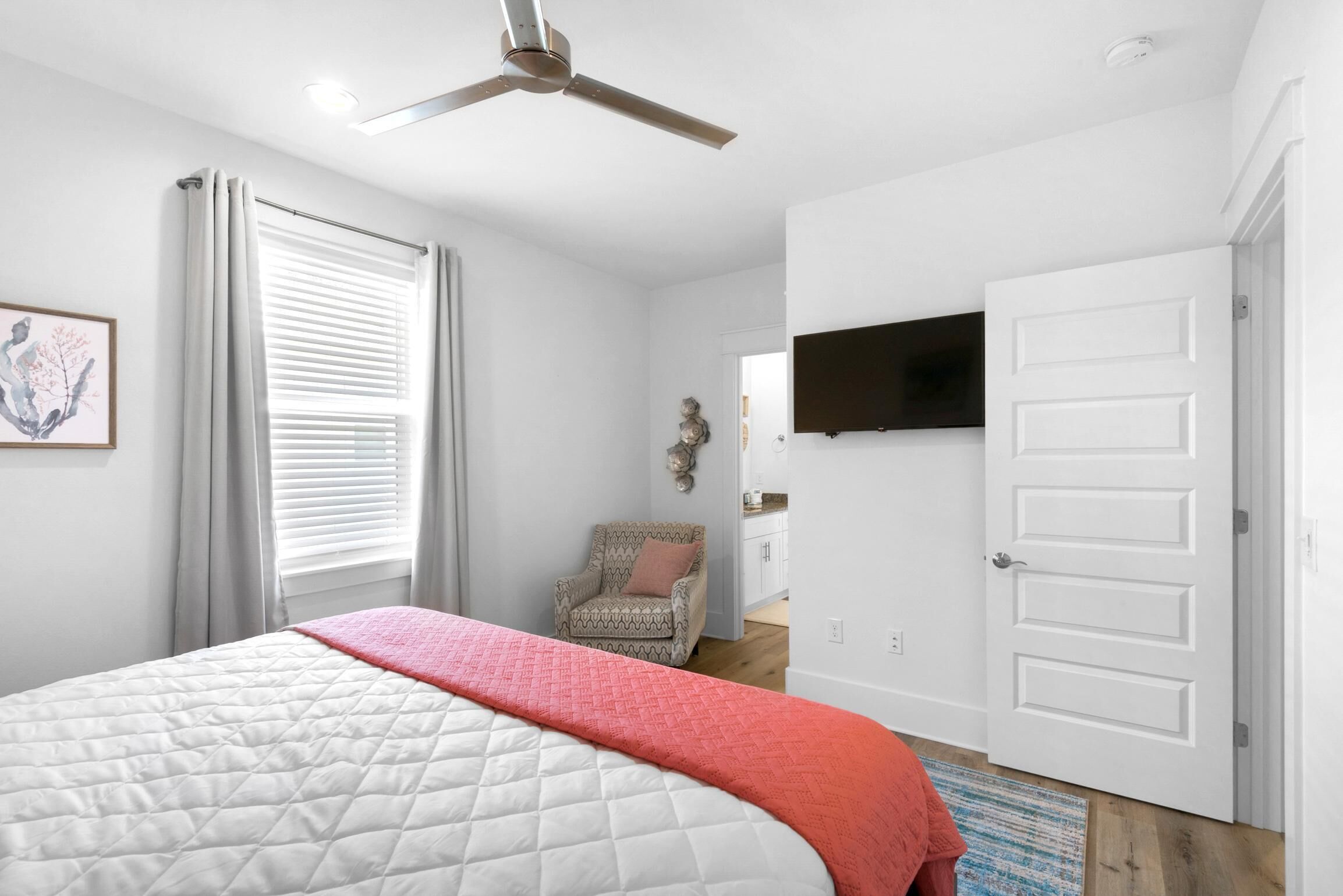 A bedroom with a bed, a ceiling fan, a TV mounted on the wall, a chair, and a door.