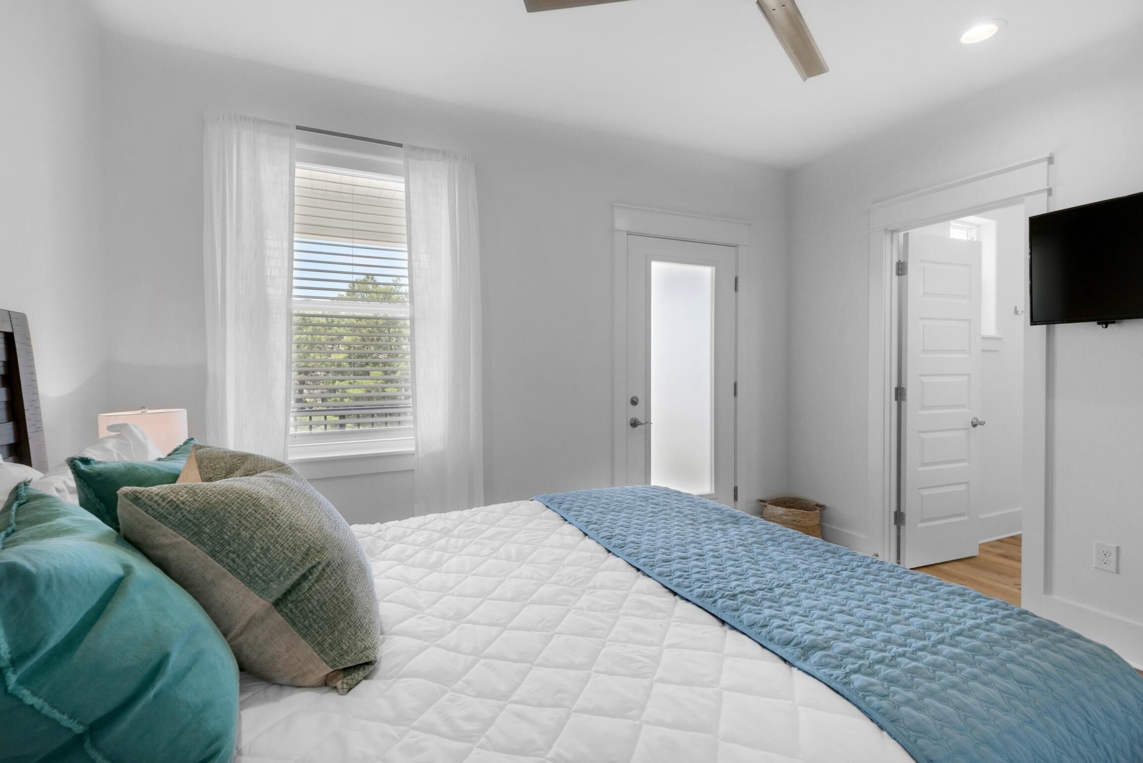 Un dormitorio con cama, una ventana con persianas, un televisor en la pared y una puerta que da a otra habitación.