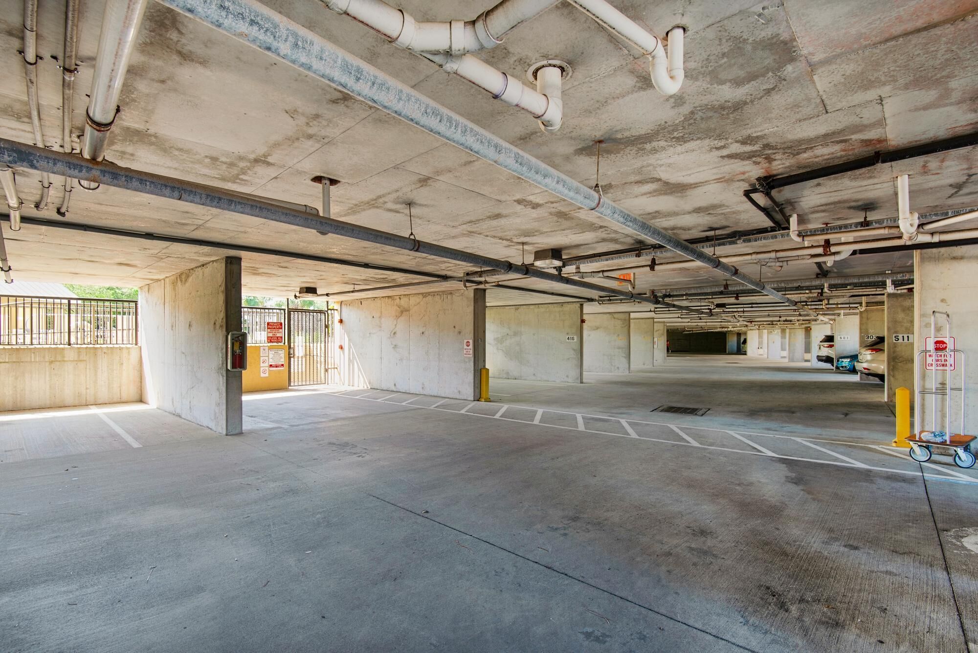 An empty parking garage with concrete floors and walls, visible pipes and ducts on the ceiling, and a gated entrance.