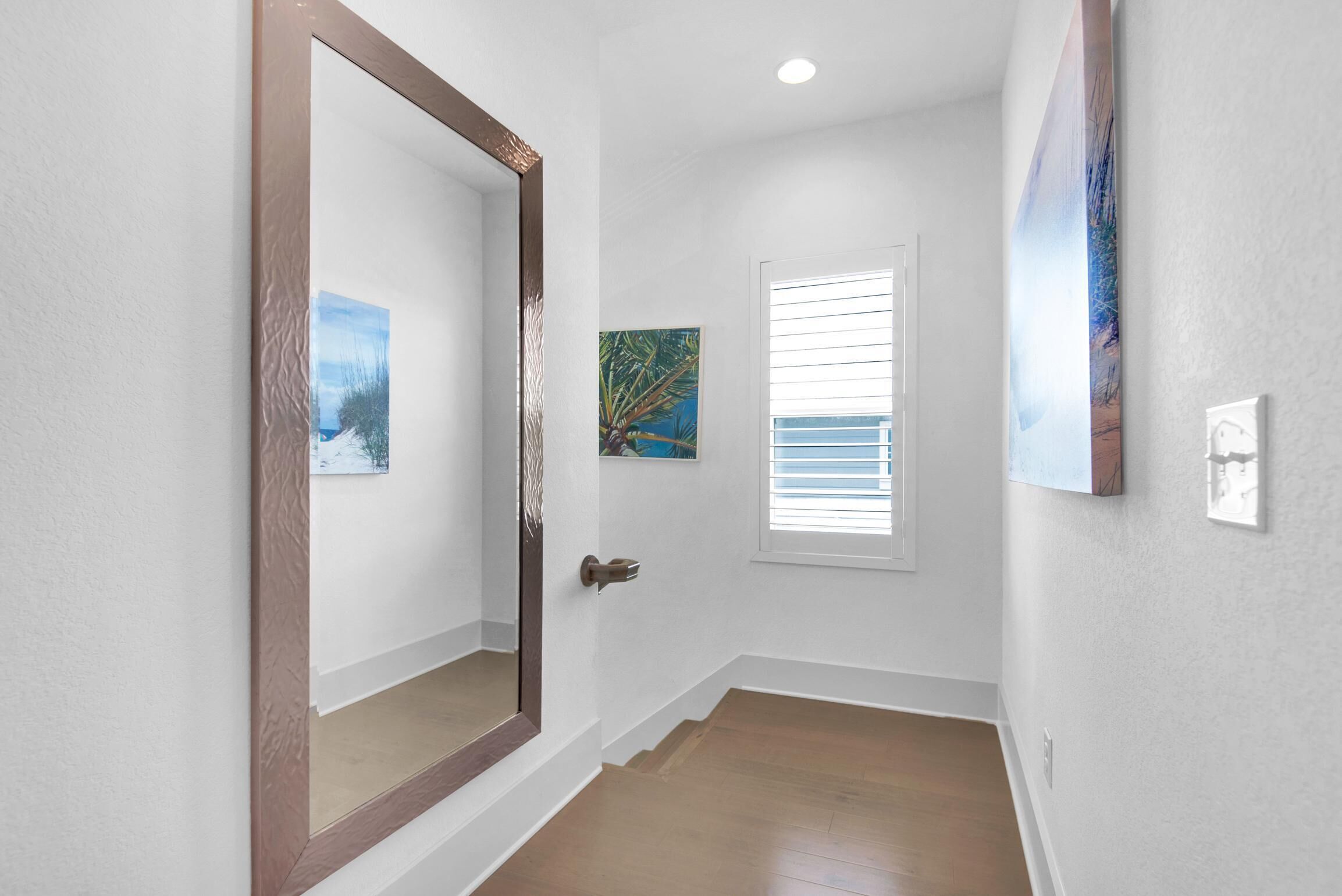 A hallway with a wooden floor, white walls, and a door with a large mirror. There are two paintings on the walls.