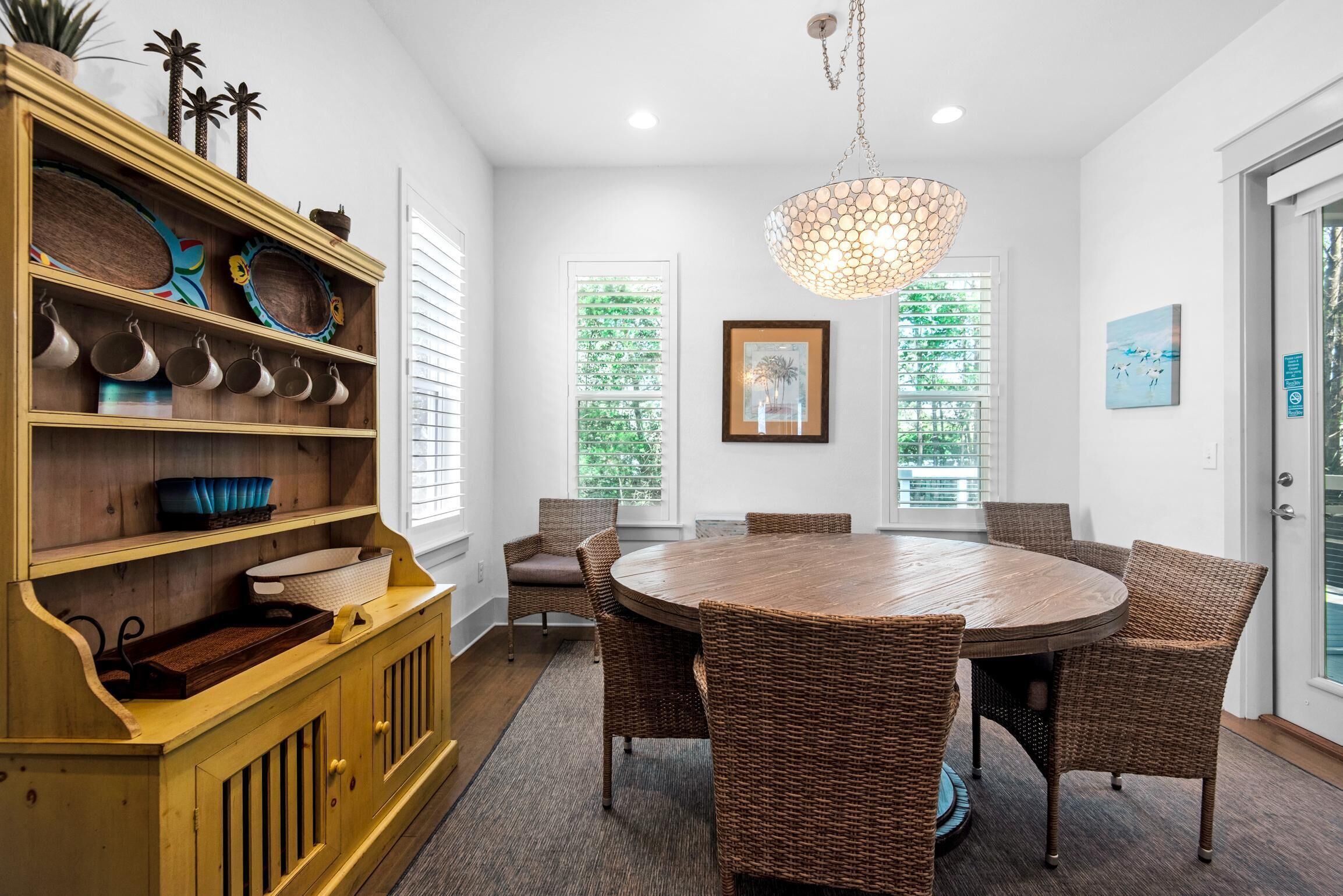 A dining area with a round wooden table surrounded by wicker chairs, a wooden cabinet with decorative items, and a hanging light fixture.