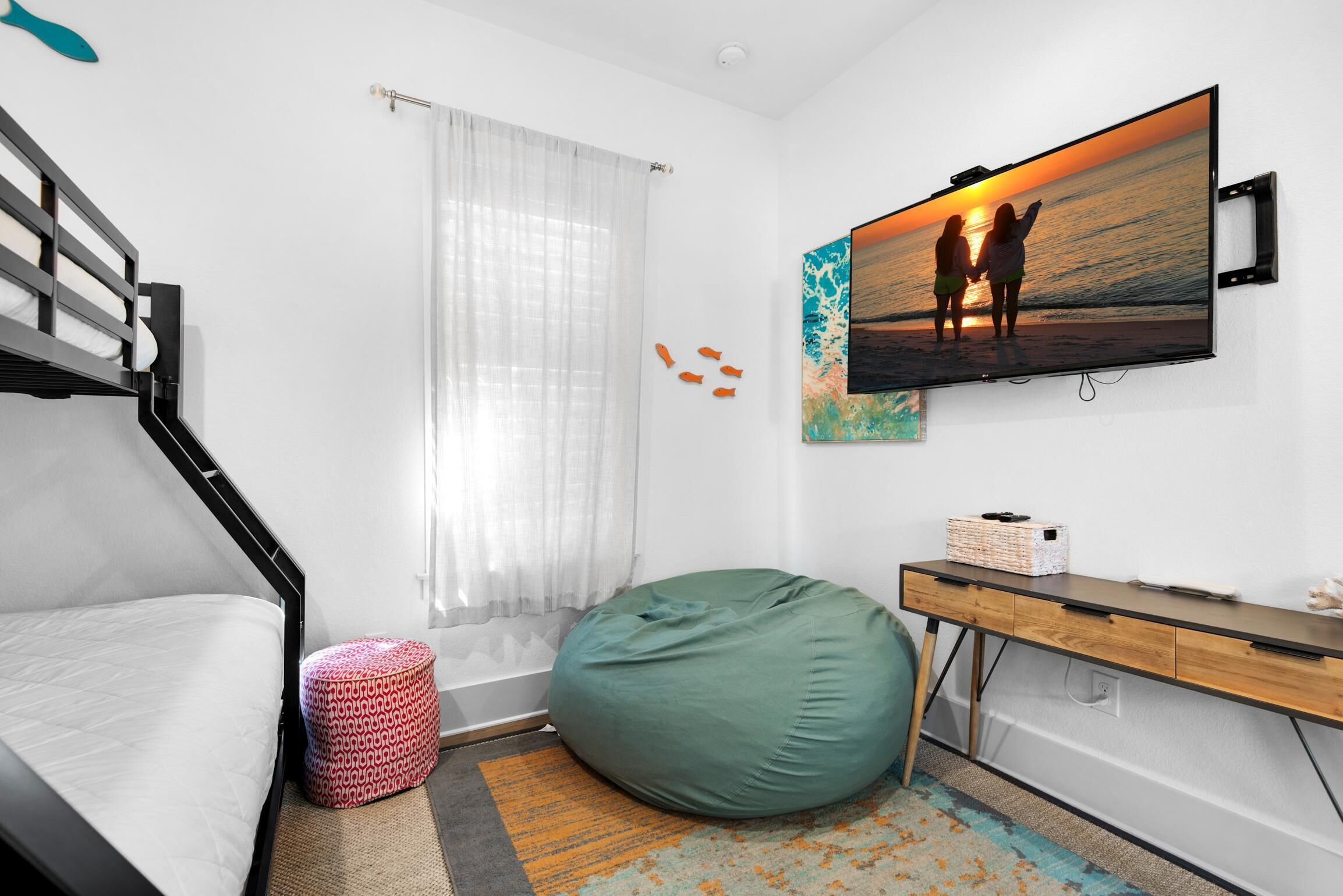 A bedroom with a bed, a desk, a bean bag, a TV on the wall, and a window with curtains.