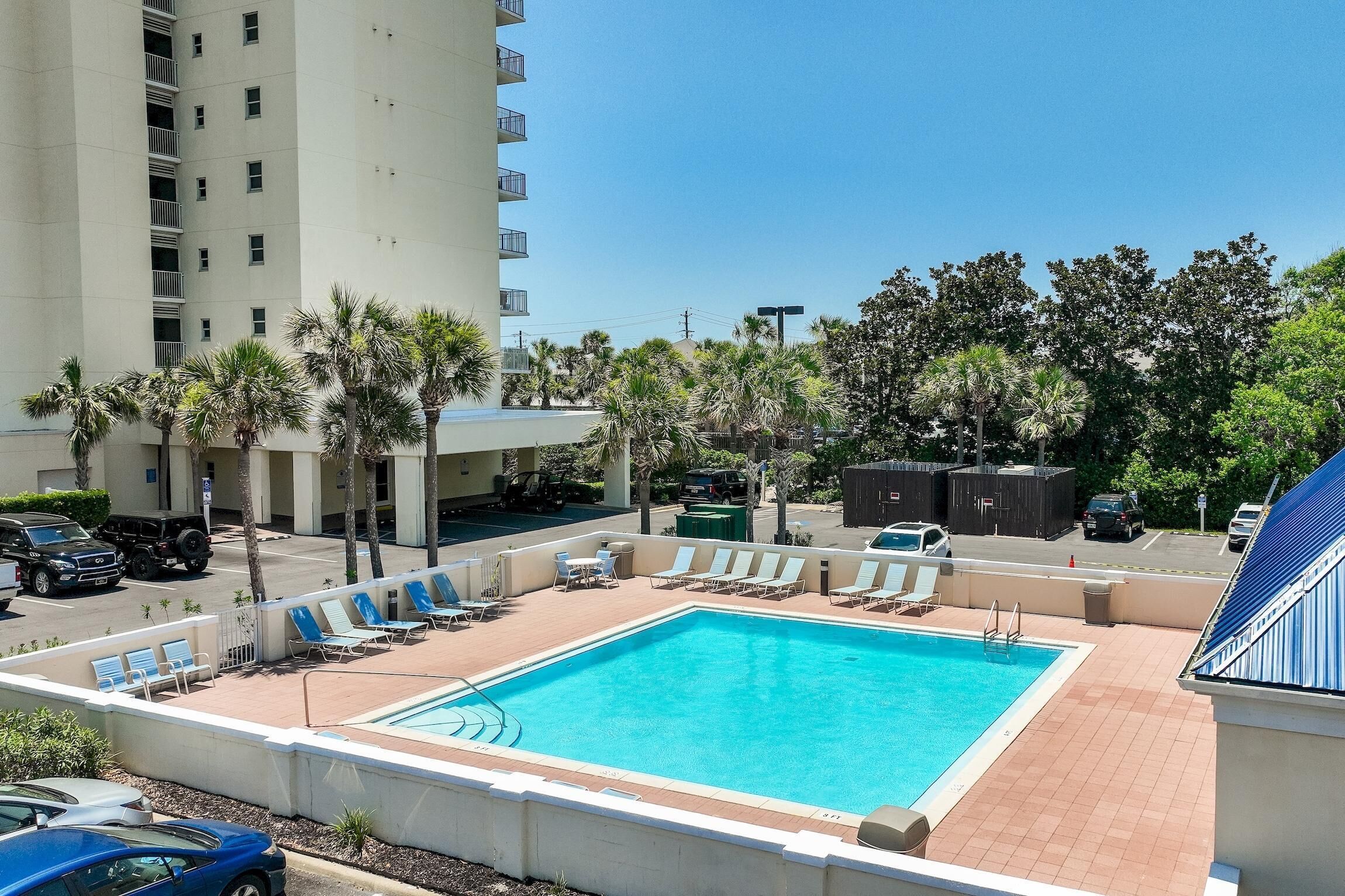 A rooftop pool with lounge chairs, surrounded by palm trees and a tall building.