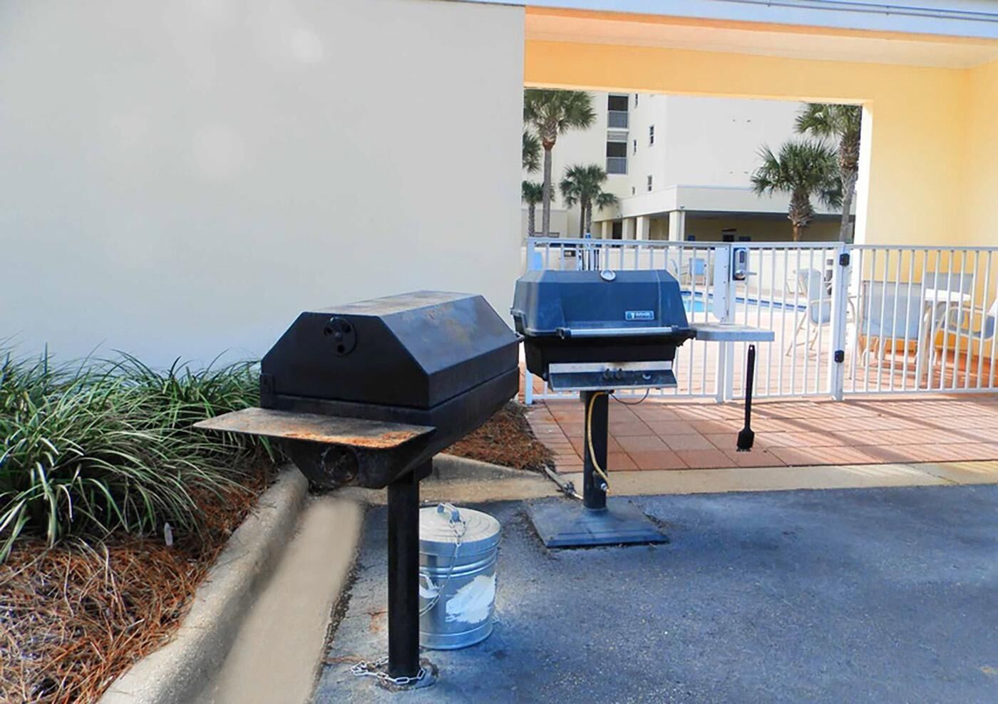 Two barbecue grills on a concrete patio with a white fence and palm trees in the background.
