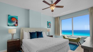 A bedroom with a large bed, a ceiling fan, artwork on the walls, a view of the ocean, and a desk with a chair. - Leeward Key 903 (Miramar Beach)