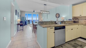A modern kitchen with light blue walls, a granite countertop, and a dishwasher. There is a dining area with a table and chairs visible in the background. - Leeward Key 903 (Miramar Beach)