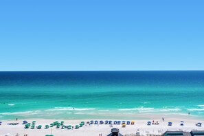 A beach with white sand and a clear blue sky. There are several umbrellas and people on the beach.