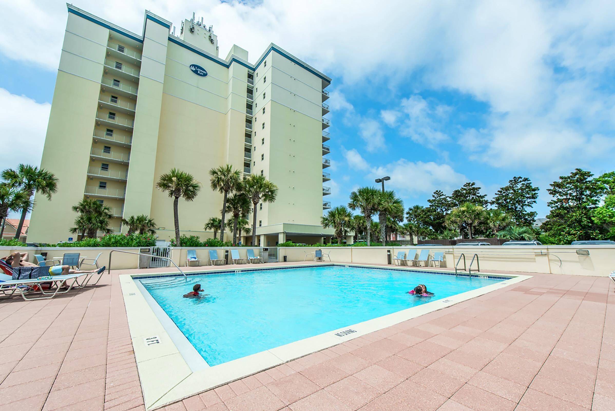 A swimming pool with two people in the water, surrounded by lounge chairs and palm trees.