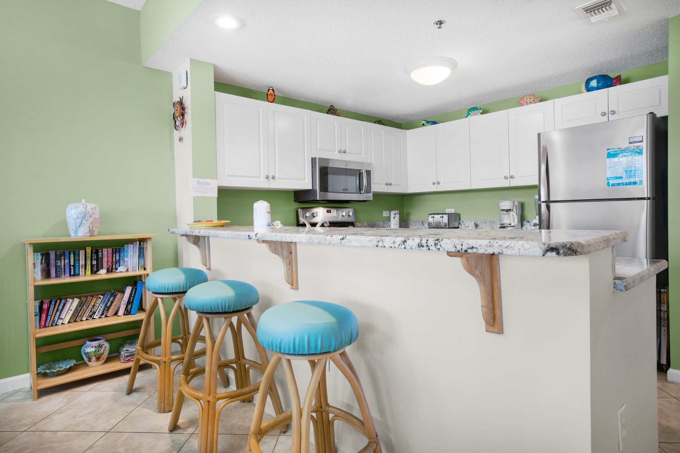 Una cocina con gabinetes blancos, microondas, refrigerador y una barra con taburetes.