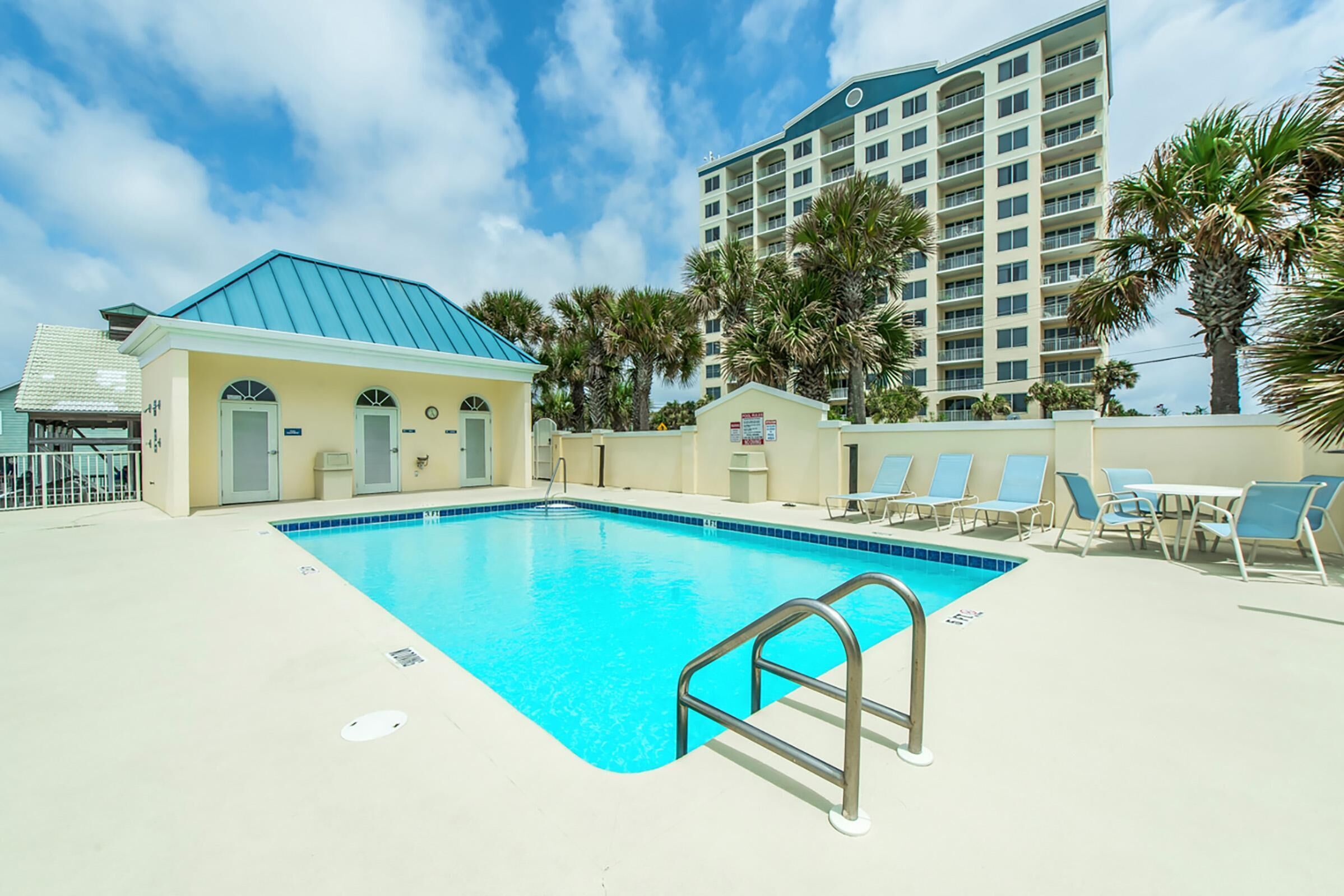A swimming pool with a metal ladder, surrounded by lounge chairs and palm trees, with a high-rise building in the background.