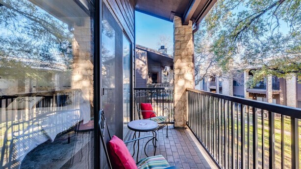 A balcony with stone pillars, a glass door, and a view of a building and trees. - Comal Condo - Riverfont Complex Across from Schlitterbahn (New Braunfels)