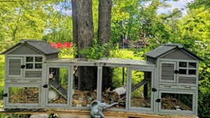 A wooden structure with two separate enclosures, each with a roof and a door, set in a garden with trees and a red chair visible in the background. - Maine river escape: free kayaks, paddle boards, tennis, cute rabbits and fun! (West Gardiner)