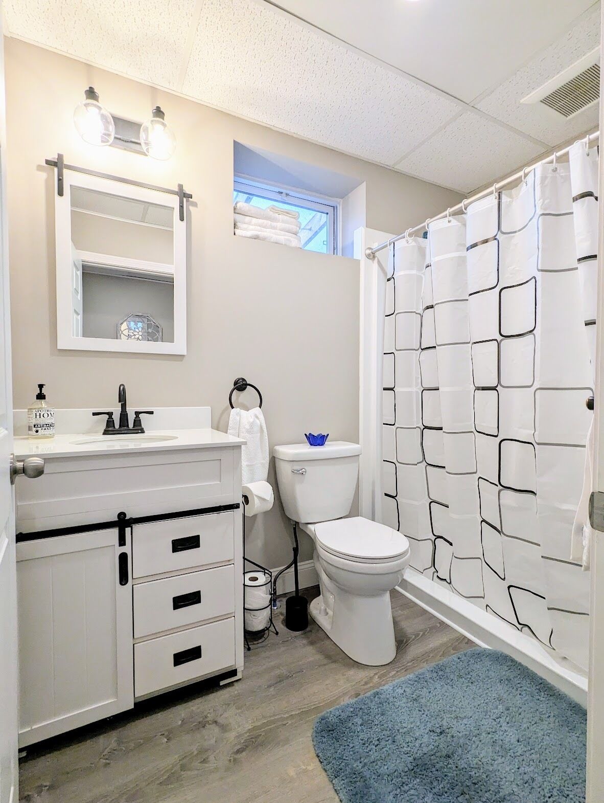 Een badkamer met een wit wastafel, toilet en douchegordijn. Er is een raam boven het toilet en een spiegel boven de wastafel.