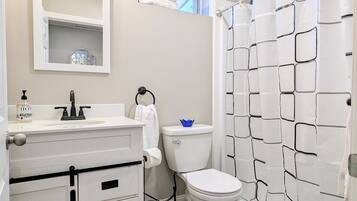 Een badkamer met een wit wastafel, toilet en douchegordijn. Er is een raam boven het toilet en een spiegel boven de wastafel.