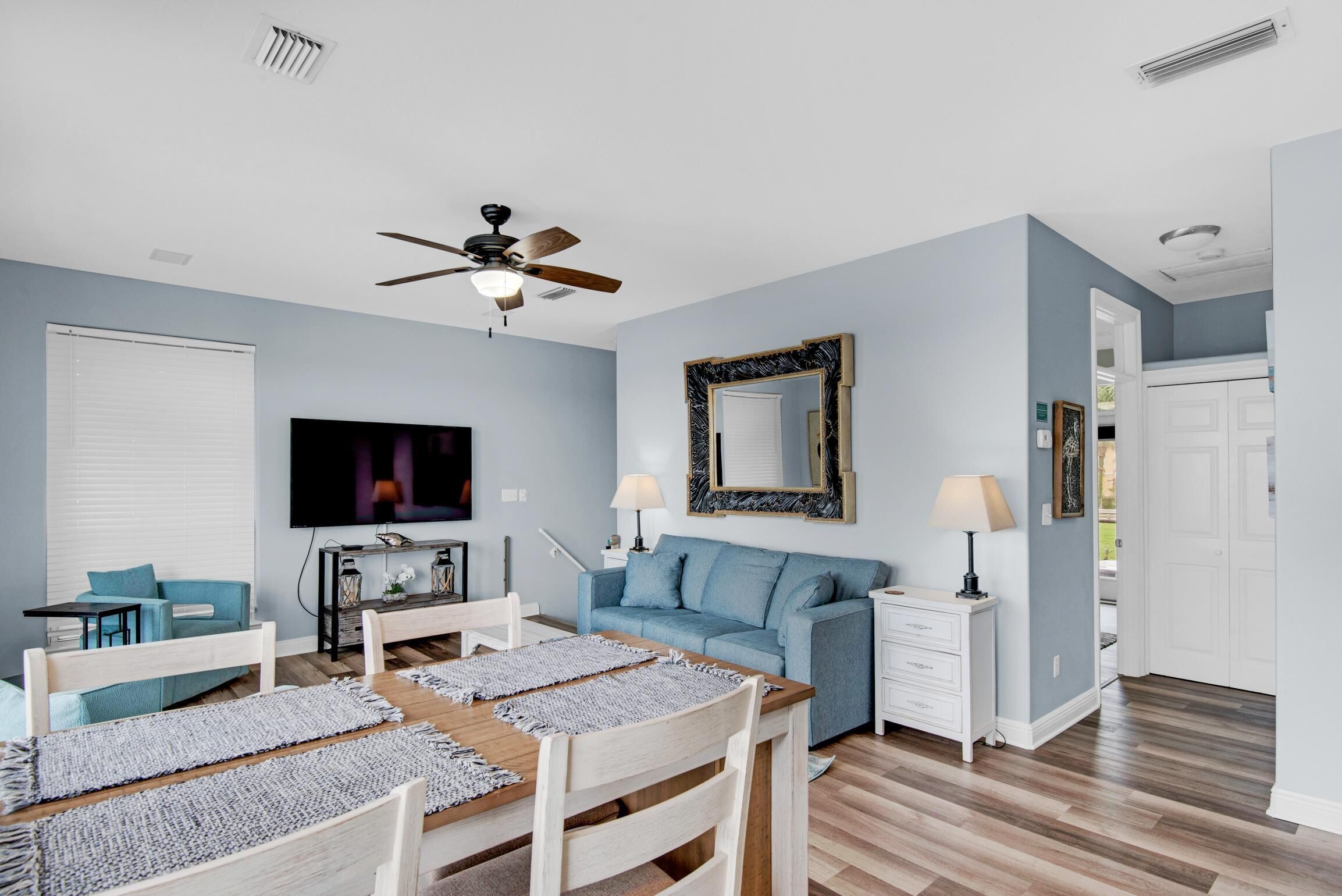 Un salon moderne doté d’un ventilateur de plafond, d’un téléviseur à écran plat, d’un canapé bleu, d’une table à manger avec des chaises et d’un meuble blanc.