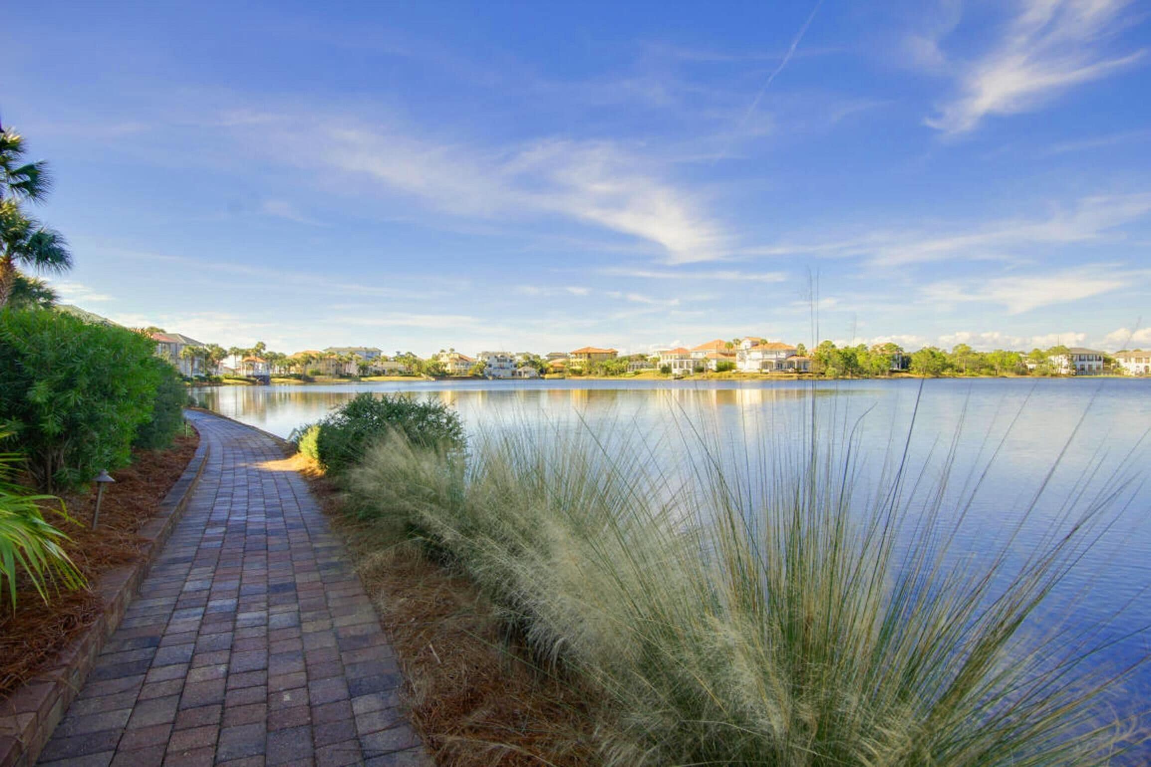 A paved pathway beside a calm lake with houses in the background.