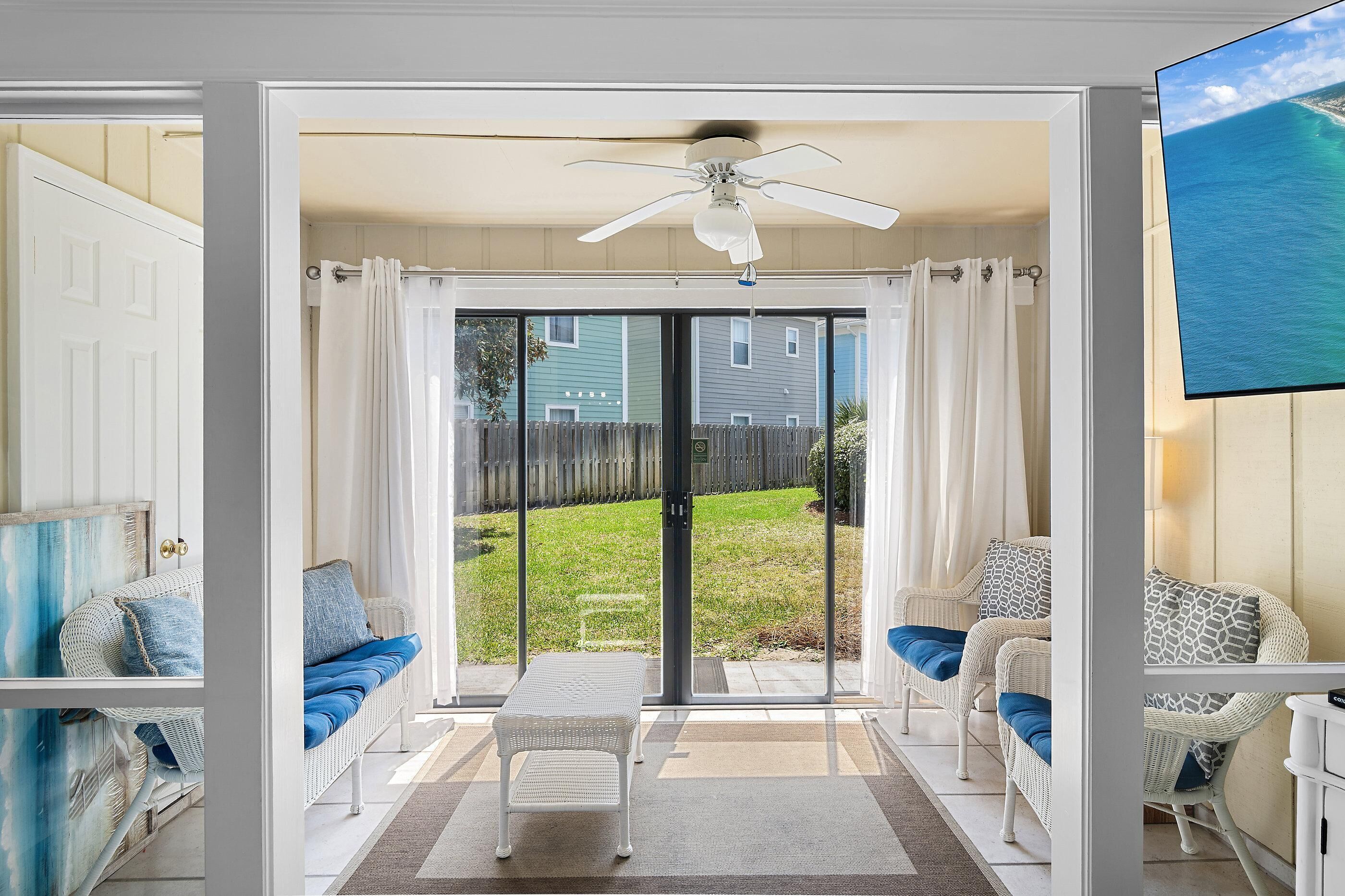 Una habitación con puerta de vidrio corrediza que da a un patio trasero, ventilador de techo y mobiliario de mimbre.