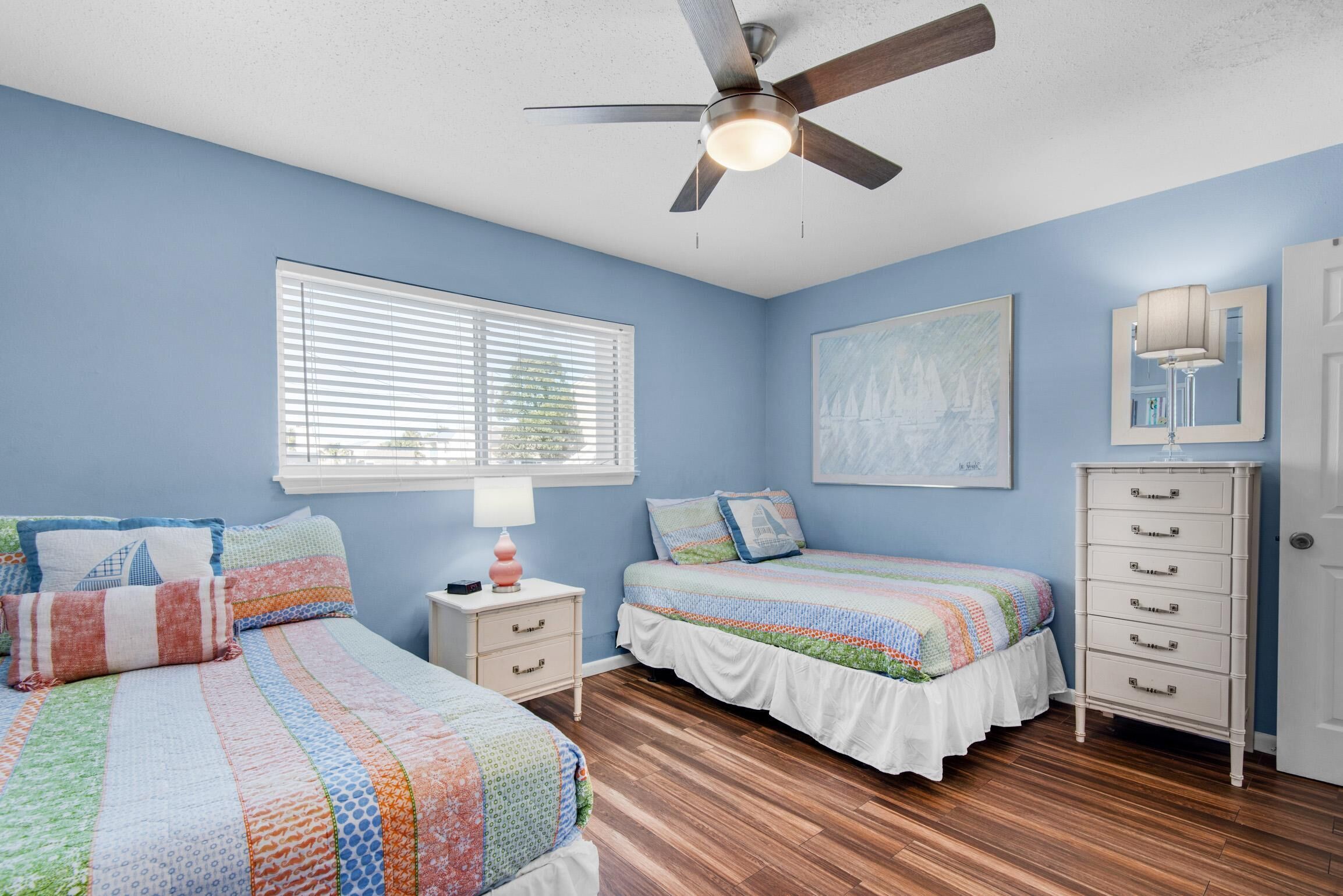 A bedroom with two beds, a ceiling fan, a window with blinds, a dresser, and a painting on the wall.
