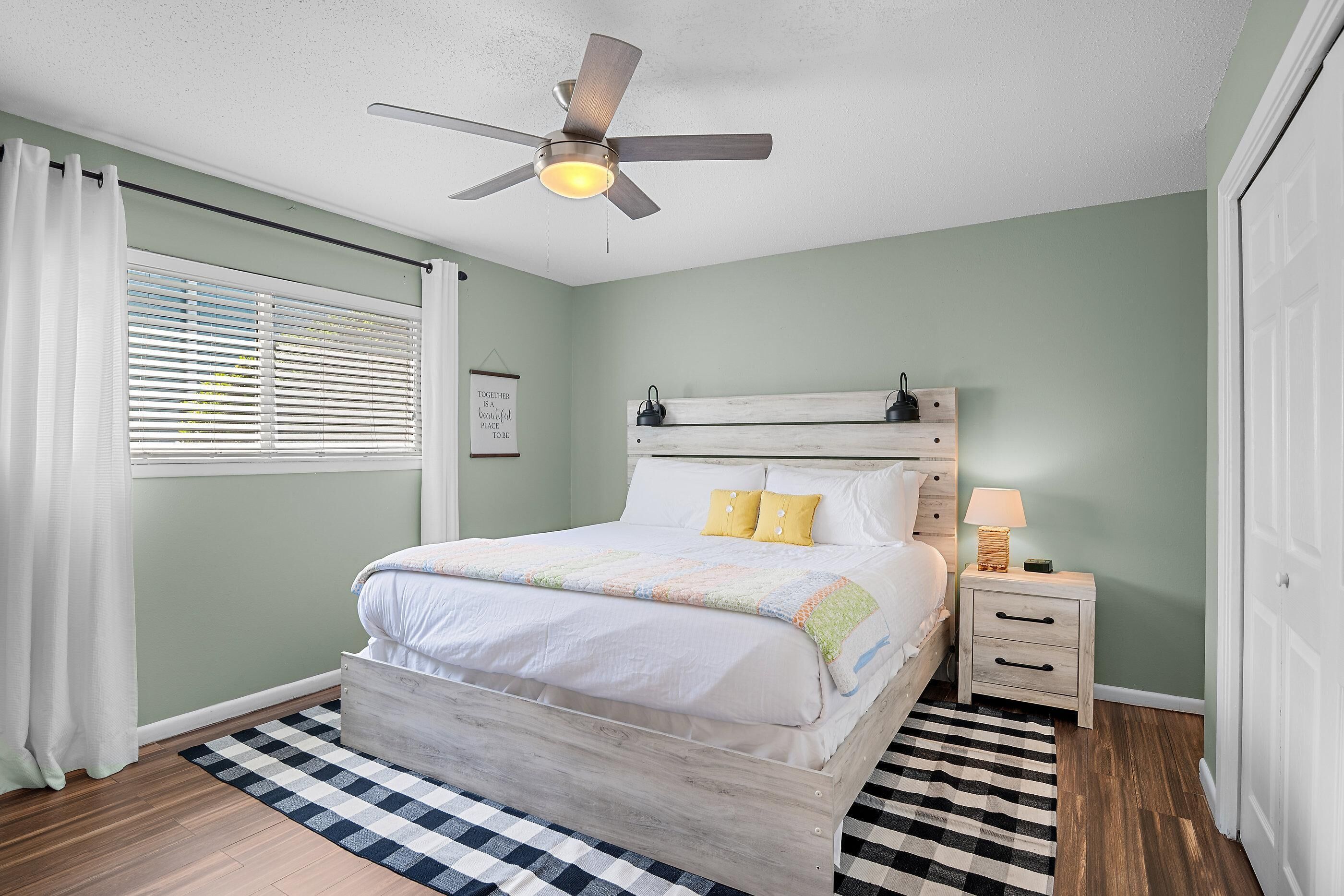 Une chambre à coucher comprenant un lit, un ventilateur de plafond, une table de chevet, une lampe et une fenêtre avec des stores.