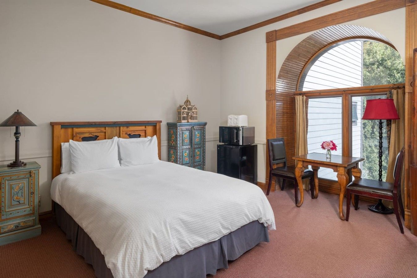 Ein Schlafzimmer mit einem großen Bett, einem Schreibtisch, einem Sessel, einer Lampe, einem Schrank und einem Fenster mit Vorhängen.