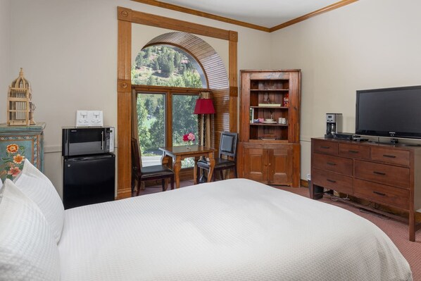 A hotel room with a large bed, a desk with a chair, a television, a wooden cabinet, and a view of trees through a window.
