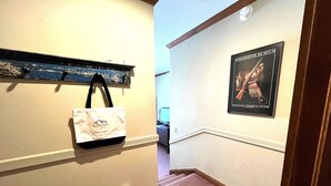 A hallway with a poster of a woman in a dress, a white tote bag hanging on the wall, and a wooden shelf above it.