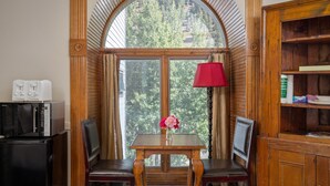 A cozy corner with a wooden table, two chairs, a red lamp, and a view of trees through a large window.