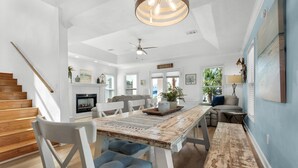 A spacious living area with a wooden staircase, a dining table with blue cushioned chairs, a fireplace, and a ceiling fan. - Another Day in Paradise (Destin)