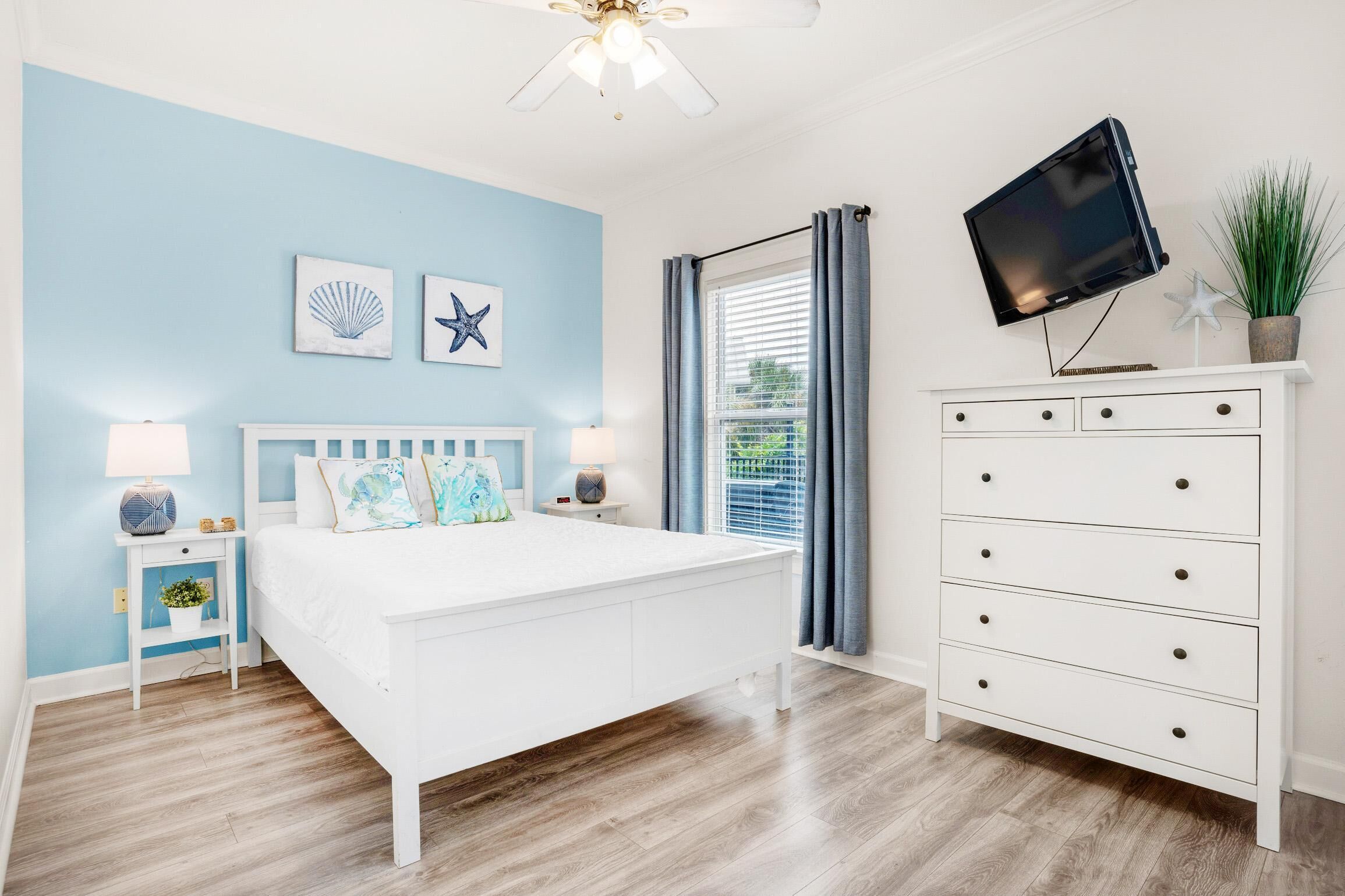 A bedroom with a bed, a dresser, a TV mounted on the wall, and a window with curtains.