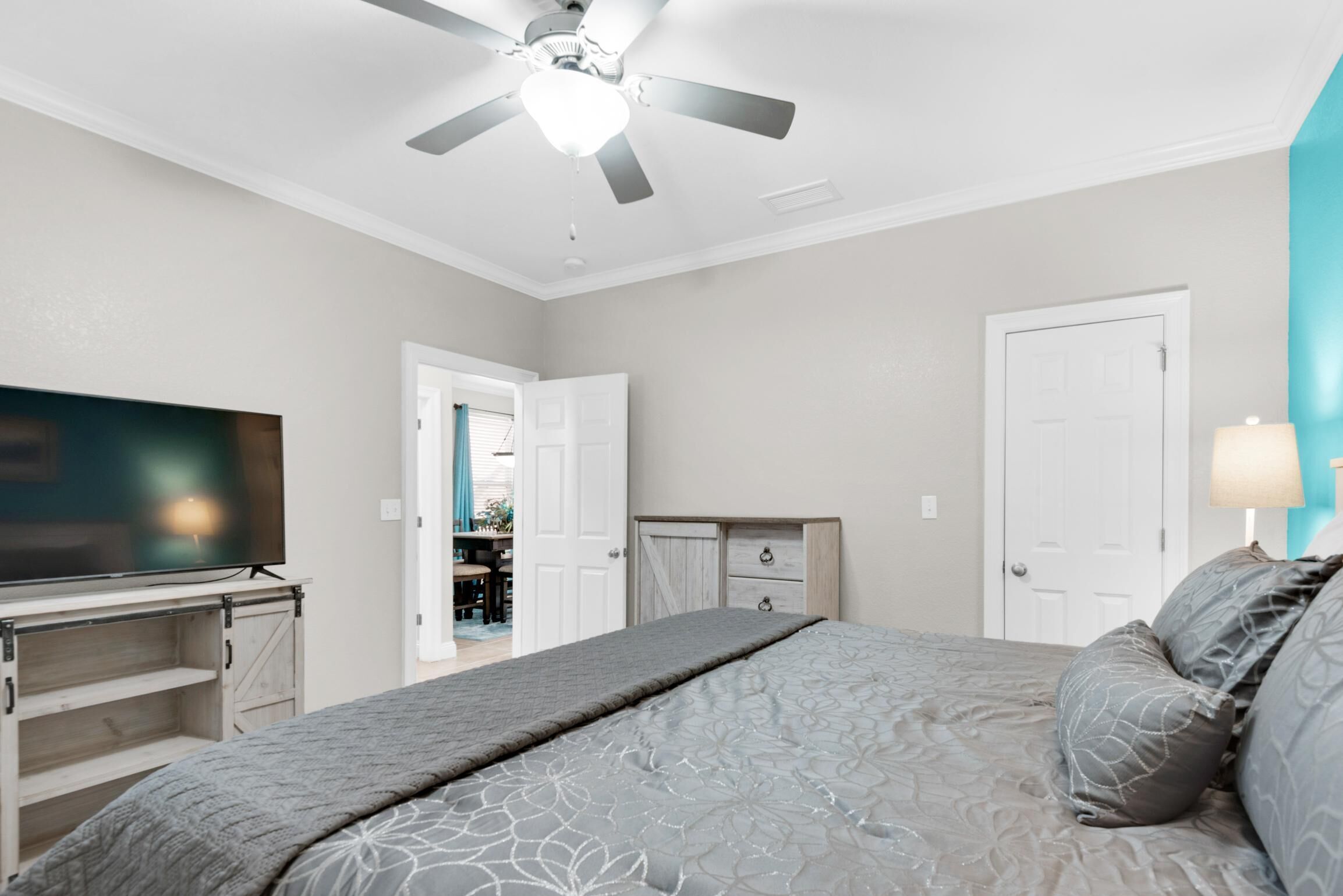 A bedroom with a bed, a TV, a ceiling fan, and a door leading to another room.