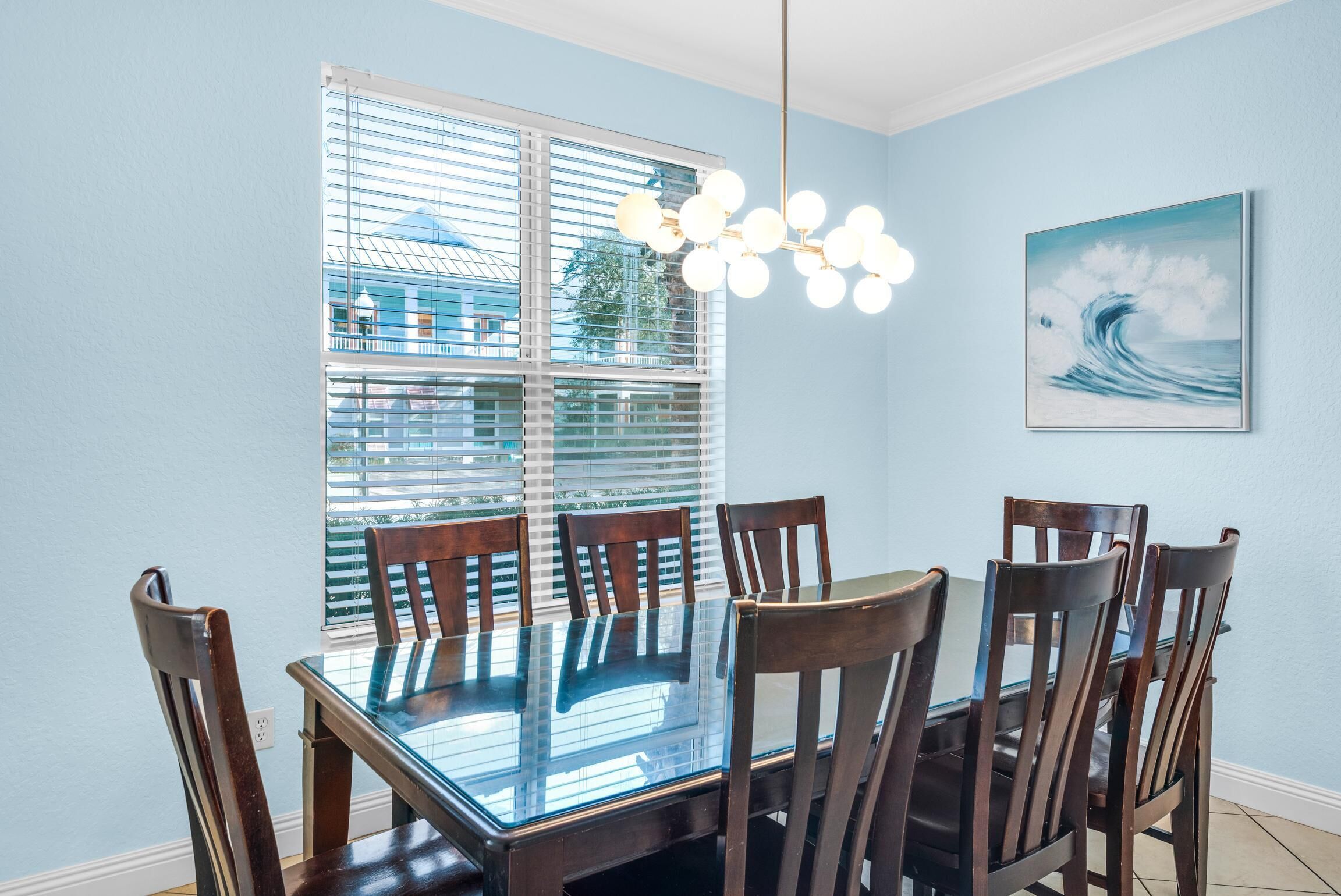 A dining area with a glass table and wooden chairs, a chandelier, and a window with blinds.