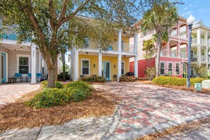 A multi-story building with a covered front porch, a brick pathway, and a tree in the foreground. - Happily Ever After (Destin)