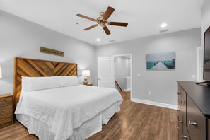 A bedroom with a wooden headboard, a bed with white bedding, a ceiling fan, a painting on the wall, and a dresser. - Vitamin Sea (Destin)