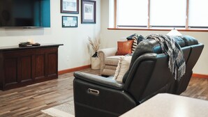 A living room with a flat-screen TV, framed pictures, a leather recliner, a sofa, and a window with natural light.