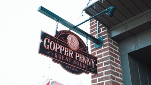 A sign for 'The Copper Penny Guest House' with a vintage design, established in 2023, hanging from a brick wall.