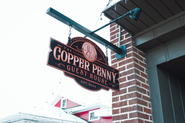 A sign for 'The Copper Penny Guest House' with a vintage design, established in 2023, hanging from a brick wall.