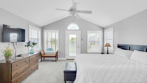 Une chambre à coucher avec un grand lit, un ventilateur de plafond, une commode, une chaise et une fenêtre avec des stores.