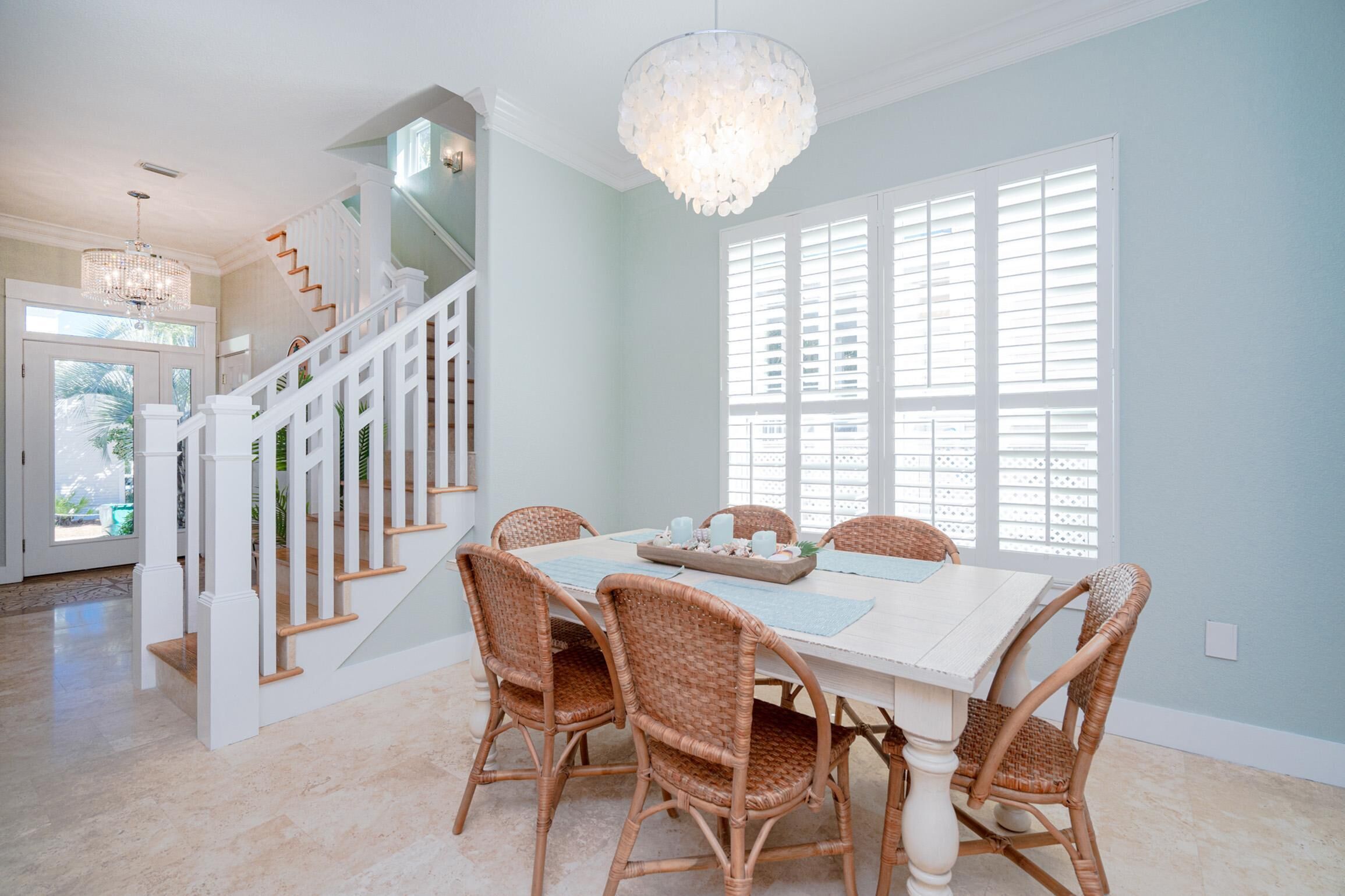 A dining area with a white table, wicker chairs, a chandelier, and a staircase with white railings.