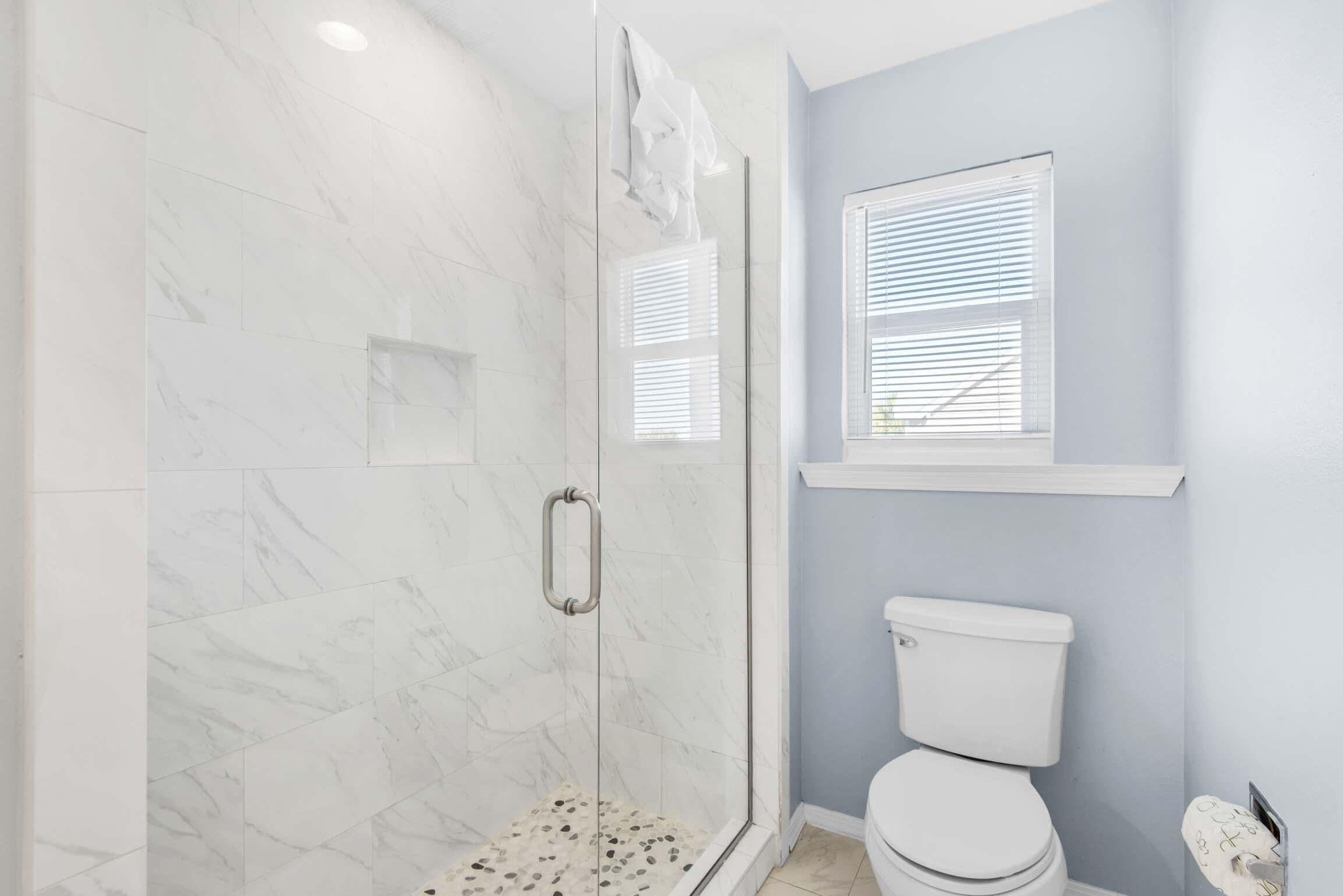 A bathroom with a glass shower enclosure, a toilet, and a window with blinds.