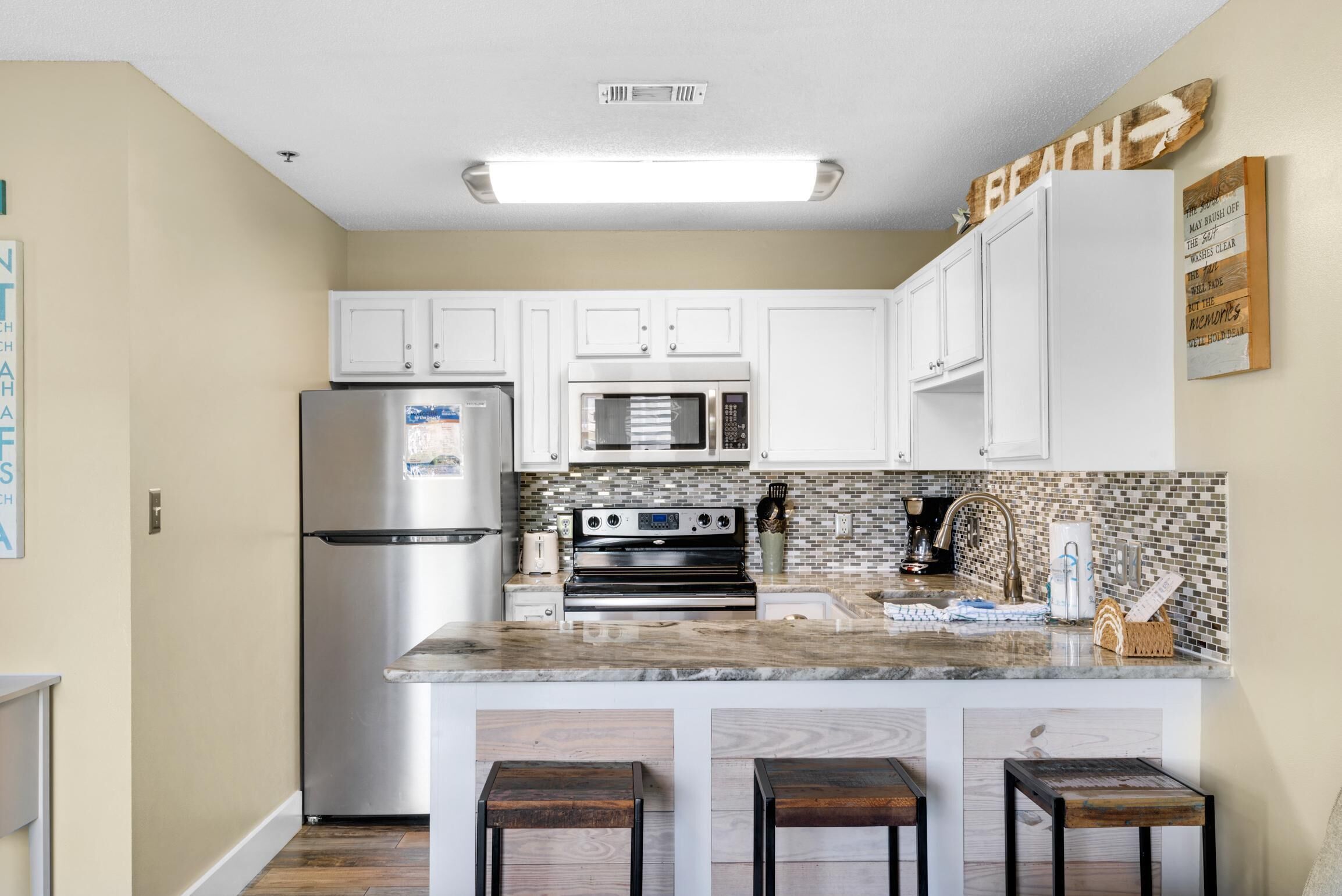 A modern kitchen with white cabinets, a stainless steel refrigerator, a microwave, an oven, and a tiled backsplash. There are three wooden stools at the island.