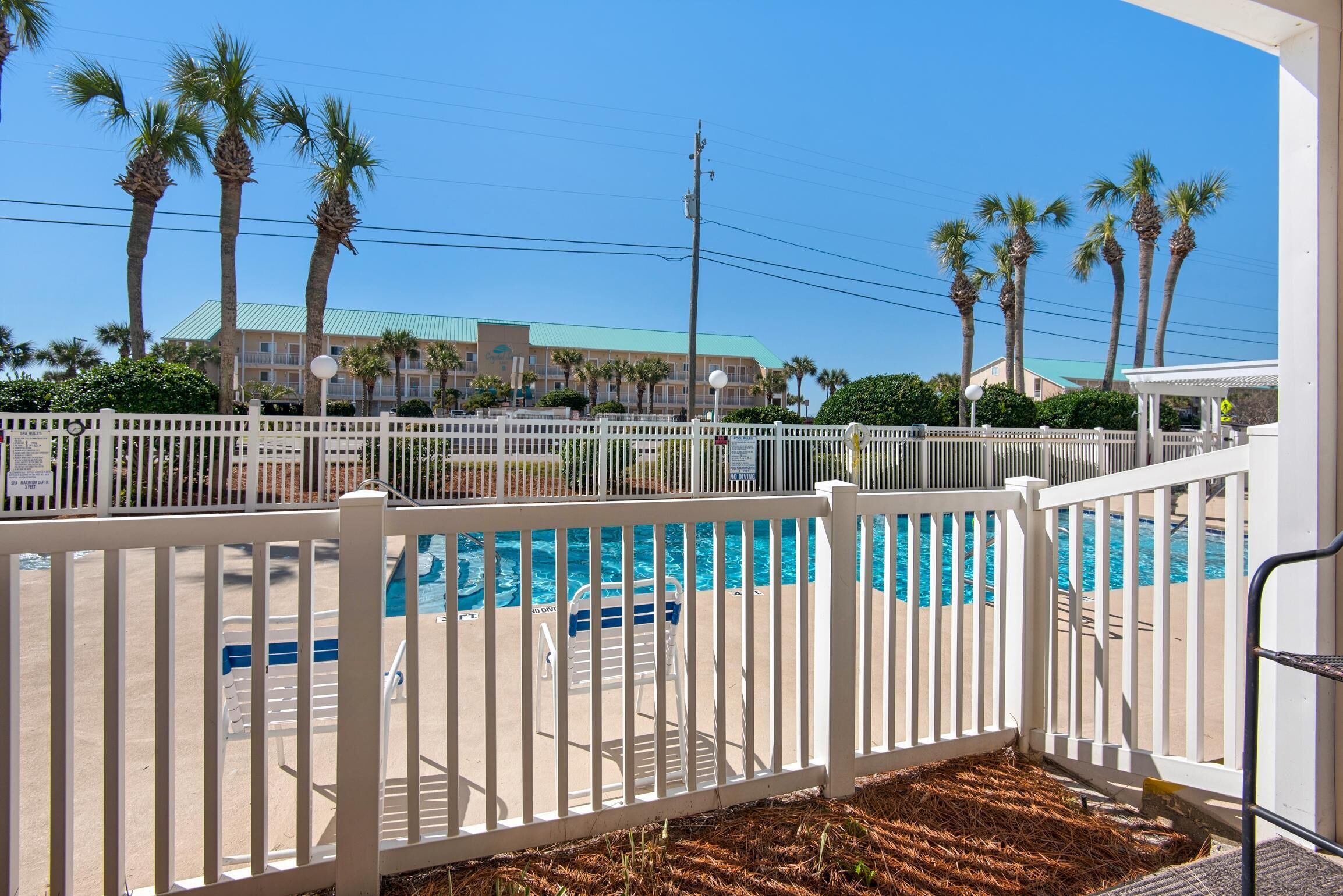 A pool area with a white fence, palm trees, and a building with a green roof in the background.